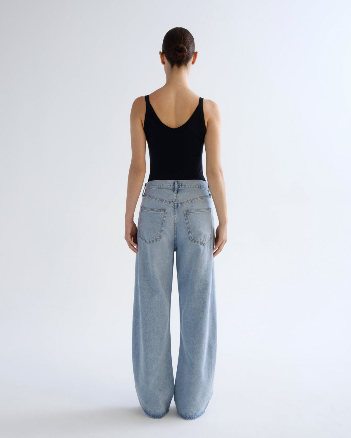 A model viewed from behind wearing a black tank top and light-wash wide-leg denim jeans against a plain white studio background.