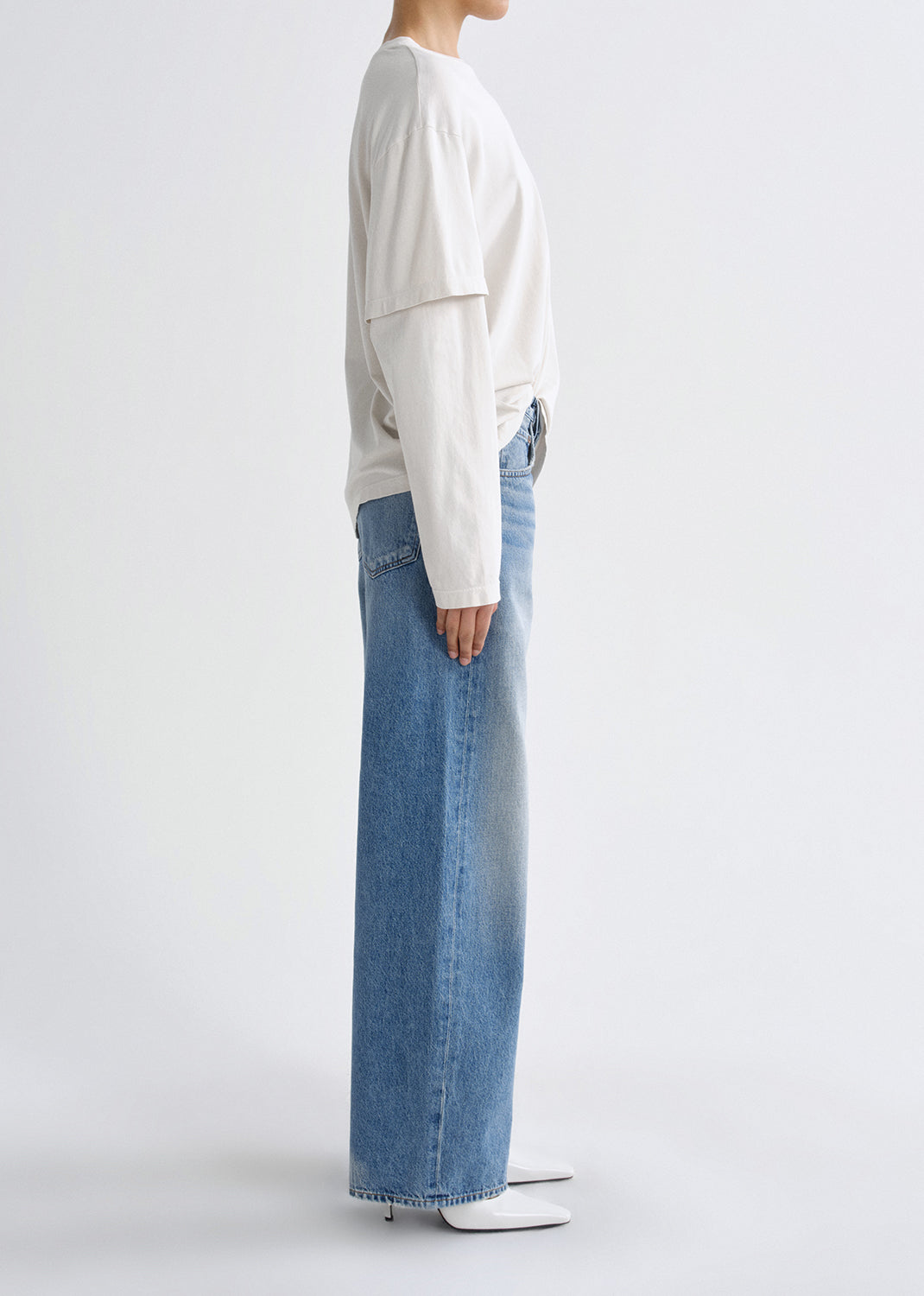 Person wearing a white long sleeve tee with light wash straight leg jeans viewed from the side on a grey background