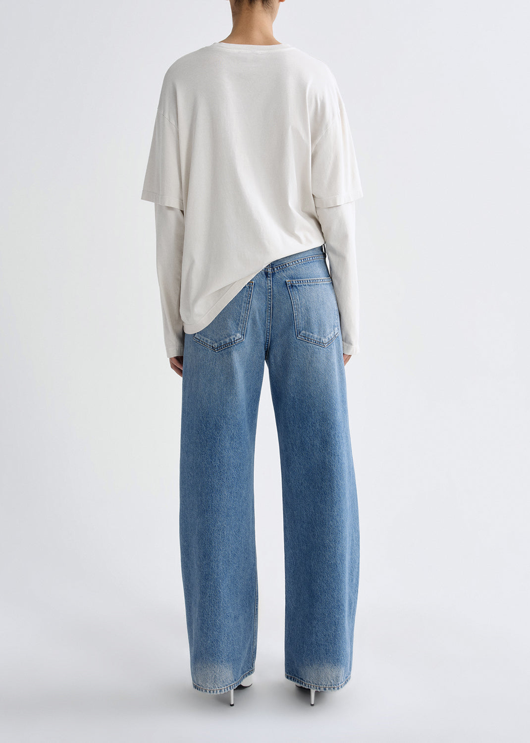 Person wearing a white long sleeve tee with light wash straight leg jeans viewed from the back on a grey background