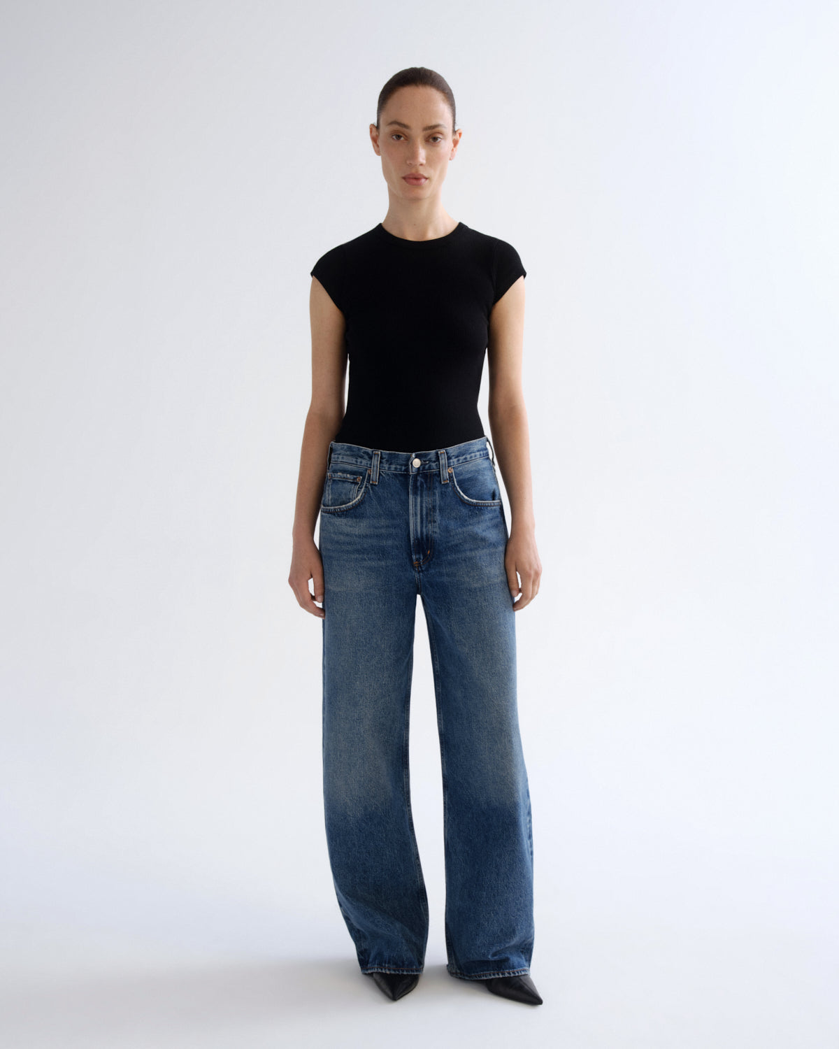 A model stands against a light gray studio background wearing a black short-sleeve top and high-waisted, wide-leg blue jeans.