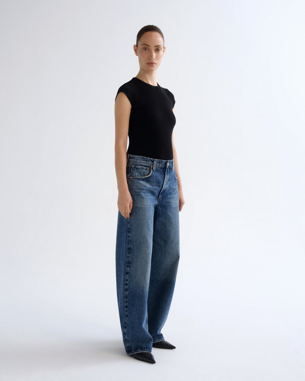 A model stands in profile wearing a black sleeveless top and wide-leg blue jeans against a clean white studio background.