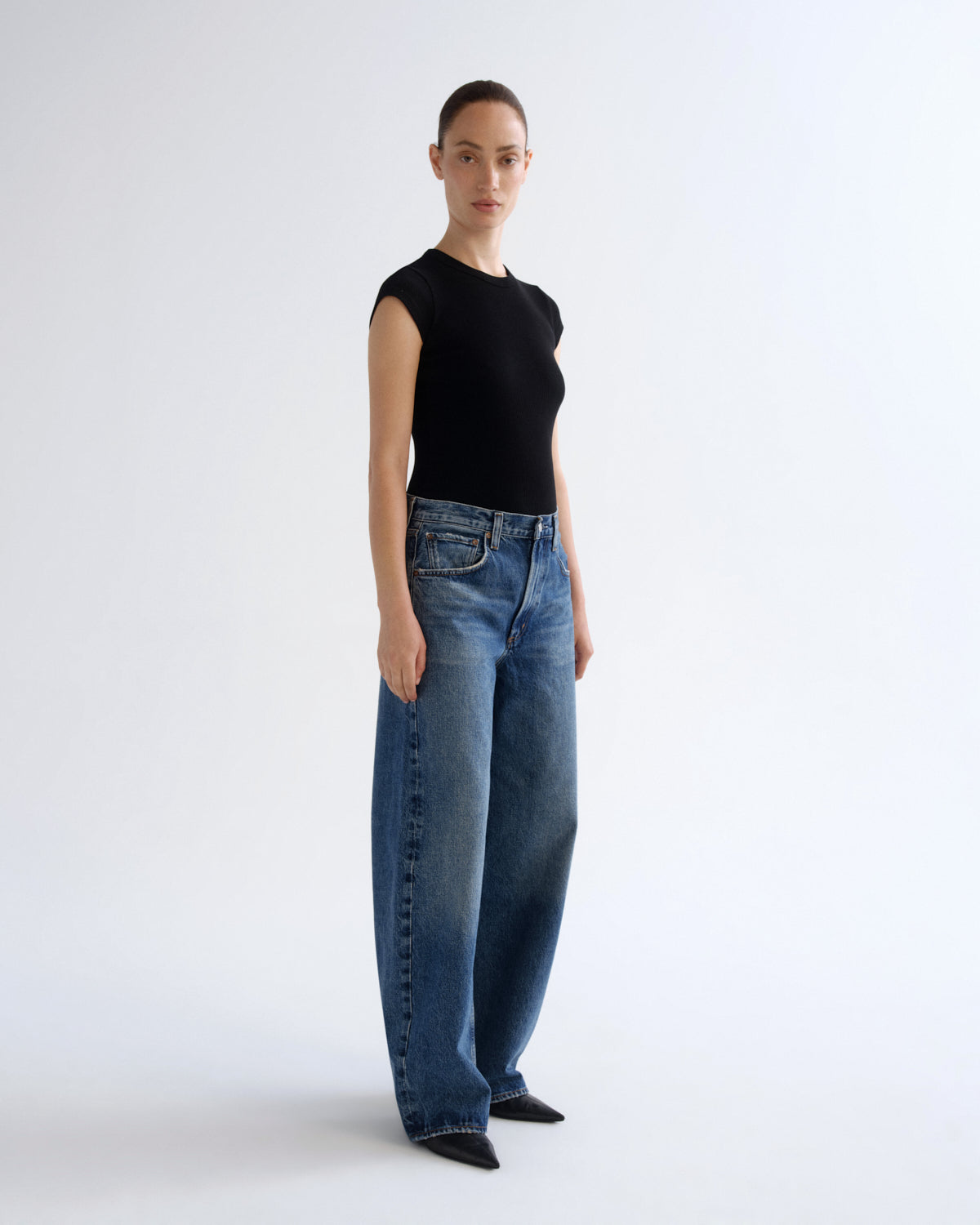 A model stands in profile wearing a black sleeveless top and wide-leg blue jeans against a clean white studio background.
