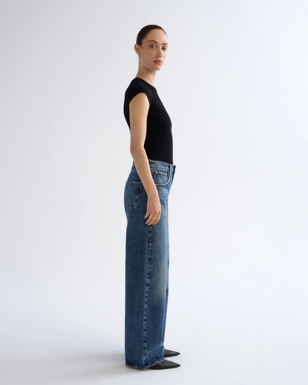A model standing in profile wearing a black short-sleeve top and wide-leg blue jeans against a clean white studio background.