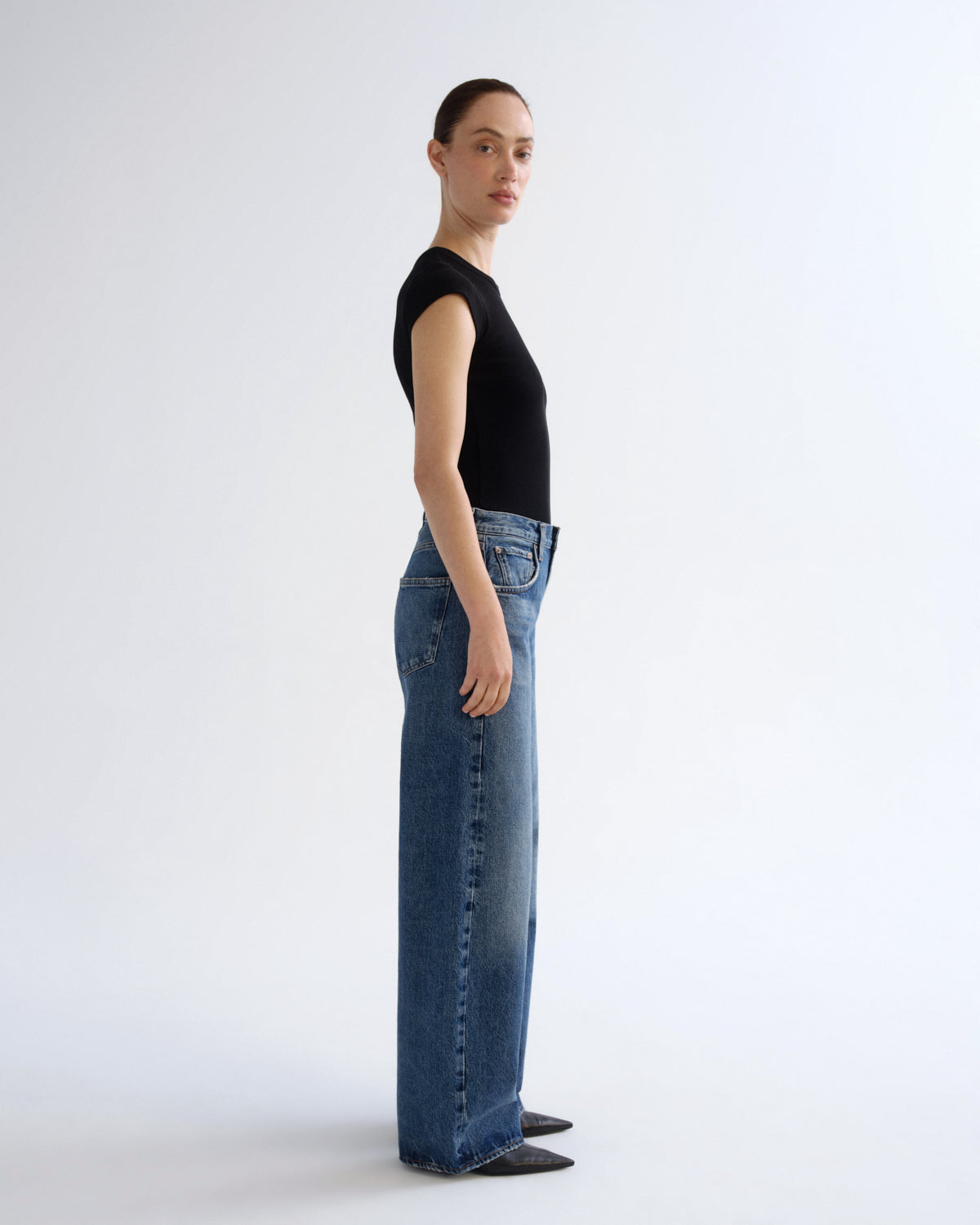 A model standing in profile wearing a black short-sleeve top and wide-leg blue jeans against a clean white studio background.