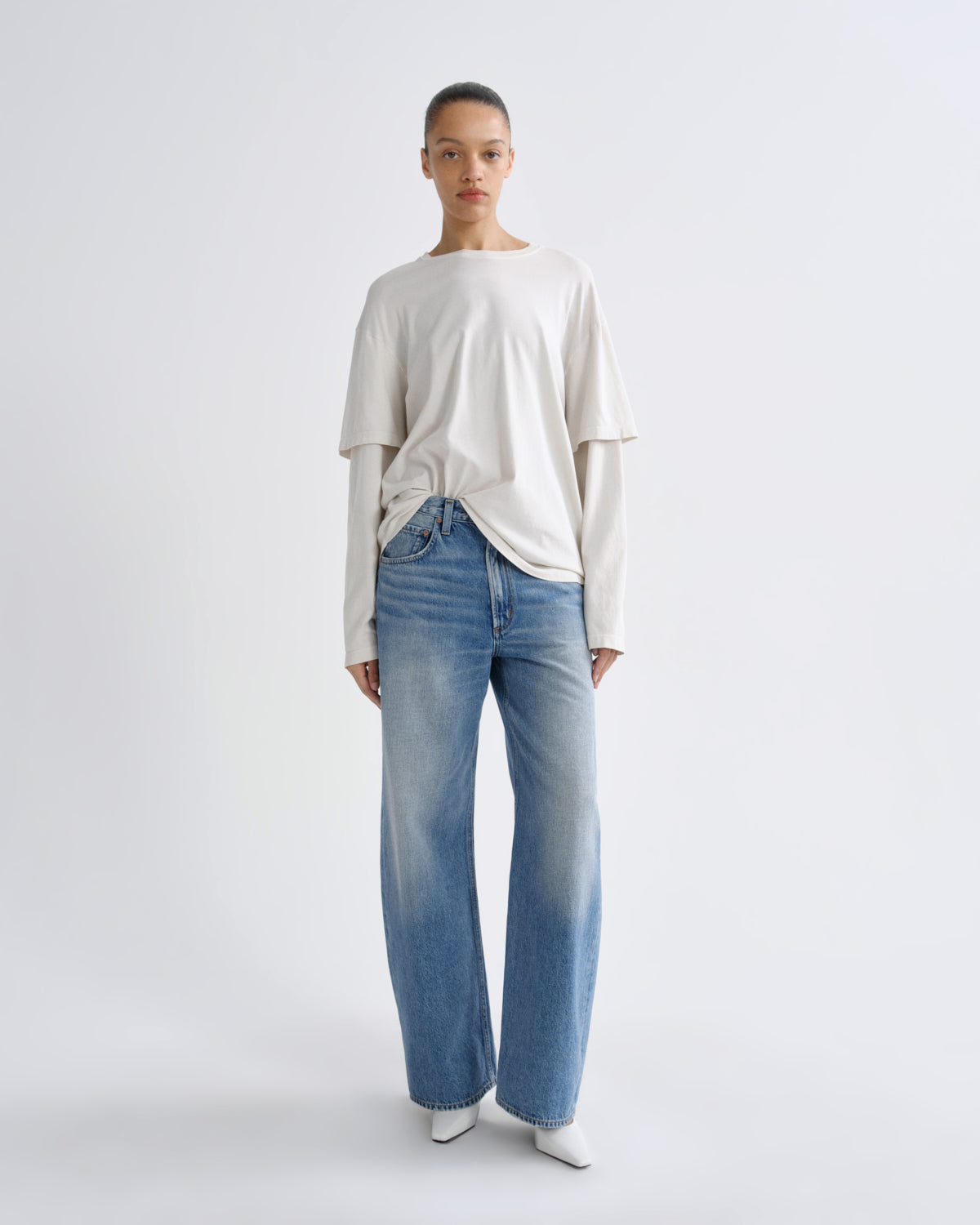 A model stands against a light gray studio background wearing a cream-colored layered long-sleeve t-shirt and light blue wide-leg jeans.