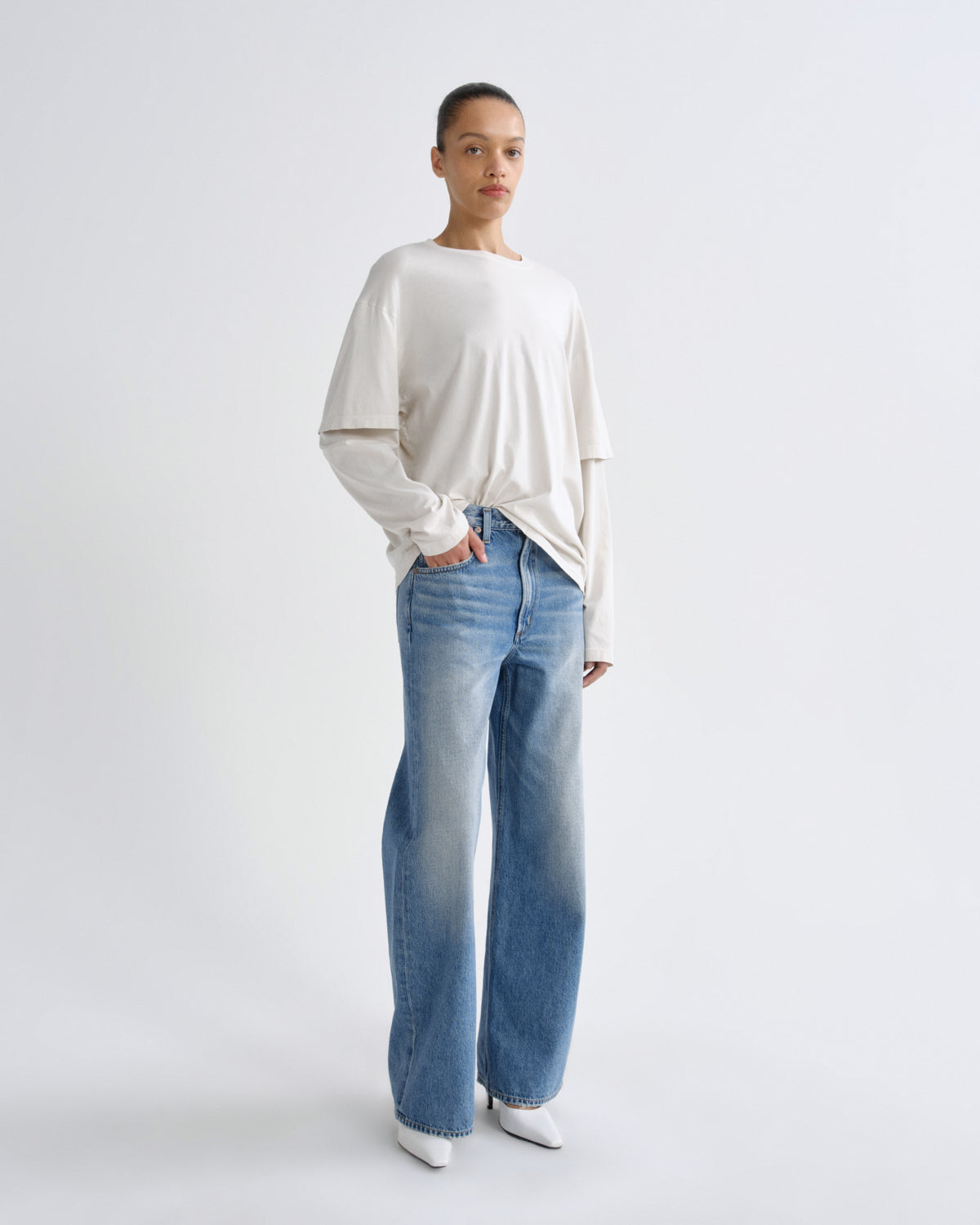 A model stands against a light gray studio background wearing a cream-colored long-sleeve t-shirt and light blue wide-leg jeans.