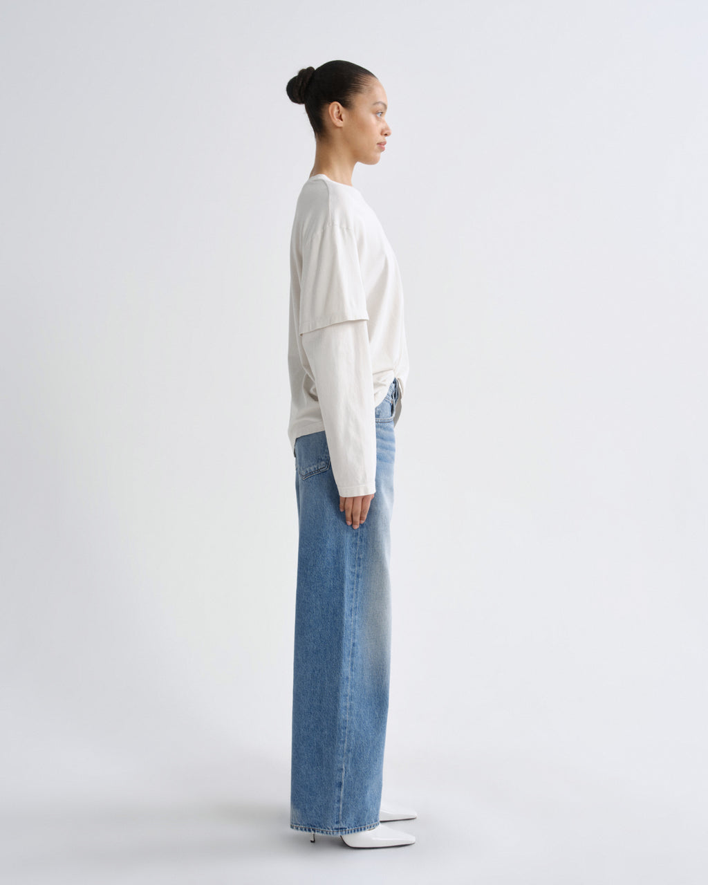A model in profile wearing a cream-colored layered long-sleeve top and wide-leg light blue jeans, standing against a plain light gray studio background.