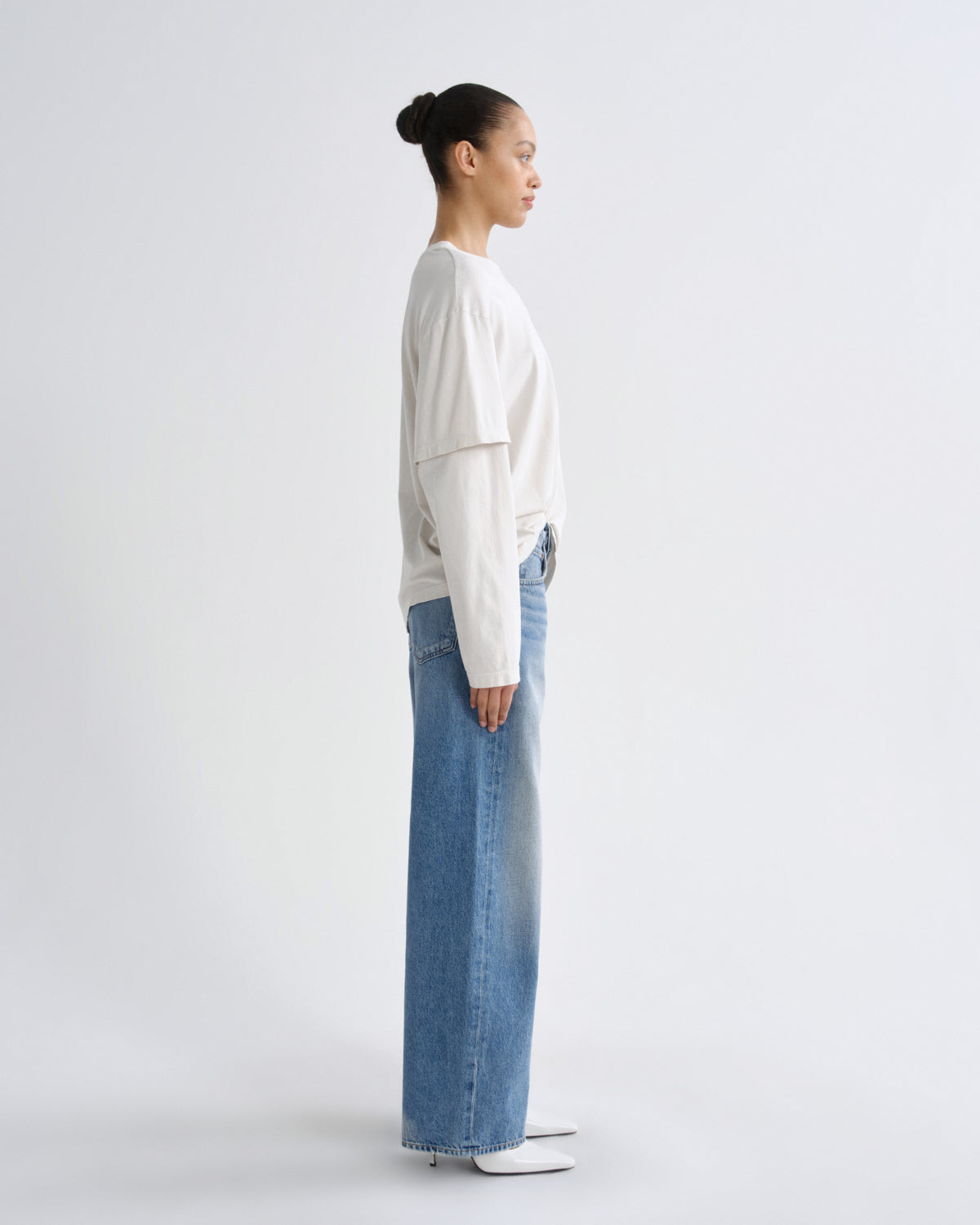 A model in profile wearing a cream-colored layered long-sleeve top and wide-leg light blue jeans, standing against a plain light gray studio background.