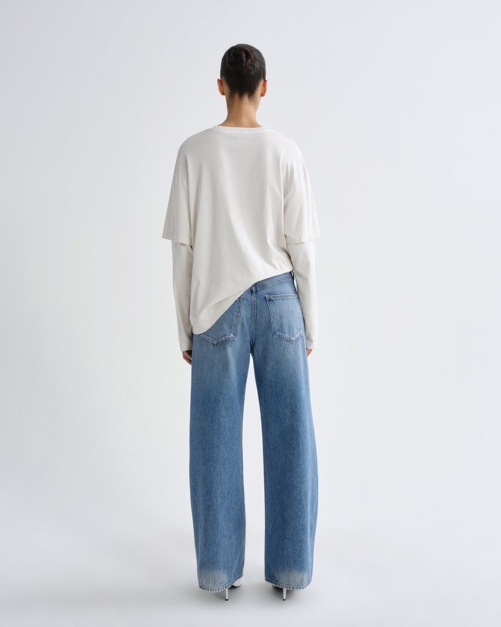 A model viewed from behind wearing a cream-colored, oversized long-sleeve top and wide-leg light blue denim jeans against a plain light gray studio background.