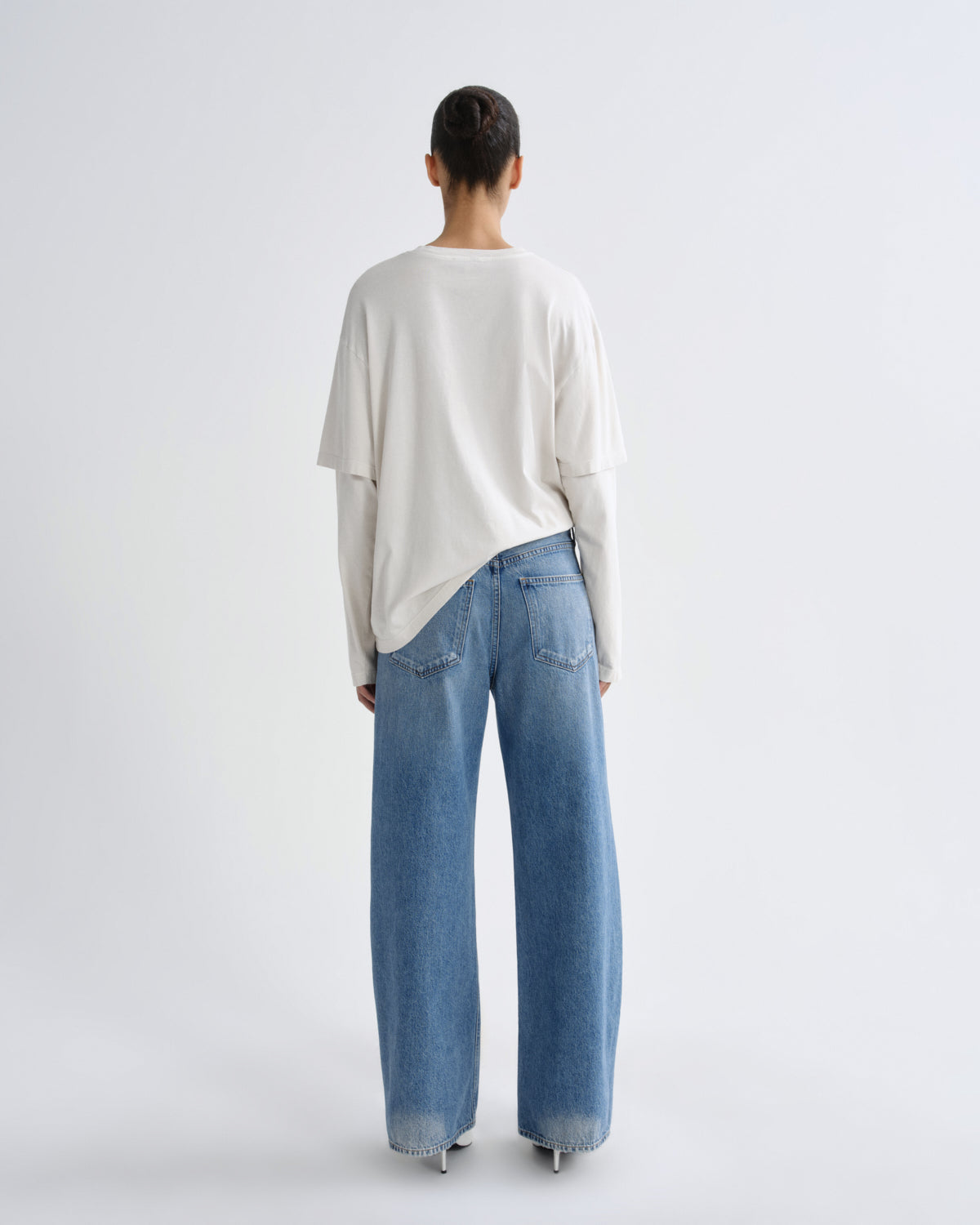 A model viewed from behind wearing a cream-colored, oversized long-sleeve top and wide-leg light blue denim jeans against a plain light gray studio background.