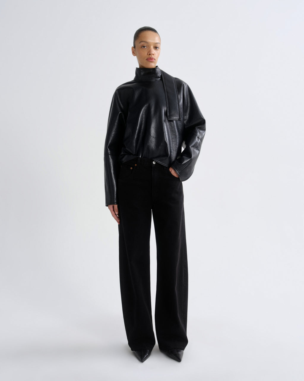 A model stands against a light gray studio background wearing a black faux-leather mock-neck jacket and black wide-leg trousers.