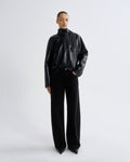 A model stands against a light gray studio background wearing a black faux-leather mock-neck jacket and black wide-leg trousers.