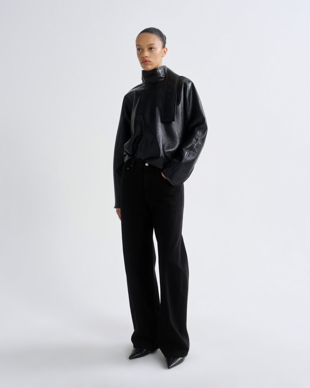 A model stands against a light gray studio background wearing a black high-neck jacket with textured panels and wide-leg black trousers.