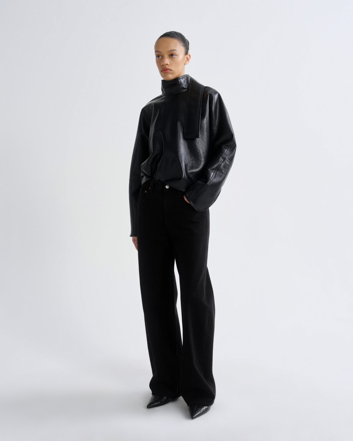 A model stands against a light gray studio background wearing a black high-neck jacket with textured panels and wide-leg black trousers.