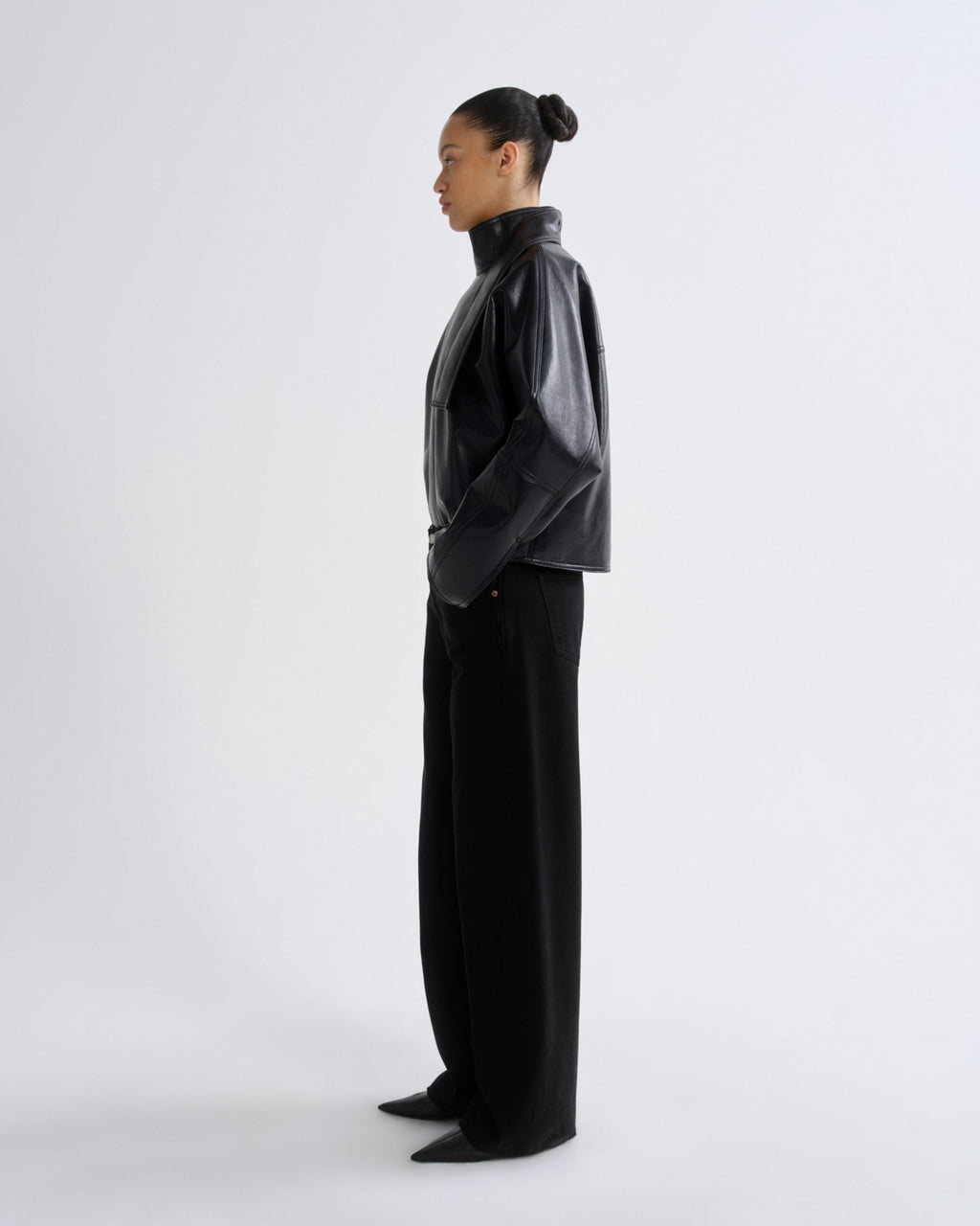 A model in side profile wearing a black leather-like cropped jacket and black wide-leg trousers, standing against a plain light gray studio background.