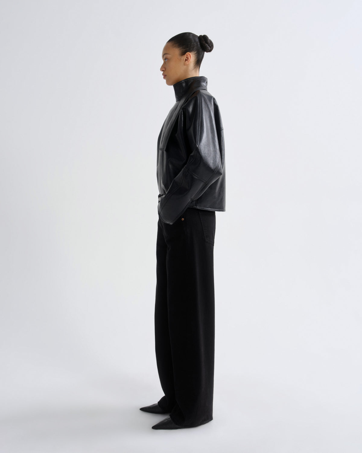 A model in side profile wearing a black leather-like cropped jacket and black wide-leg trousers, standing against a plain light gray studio background.