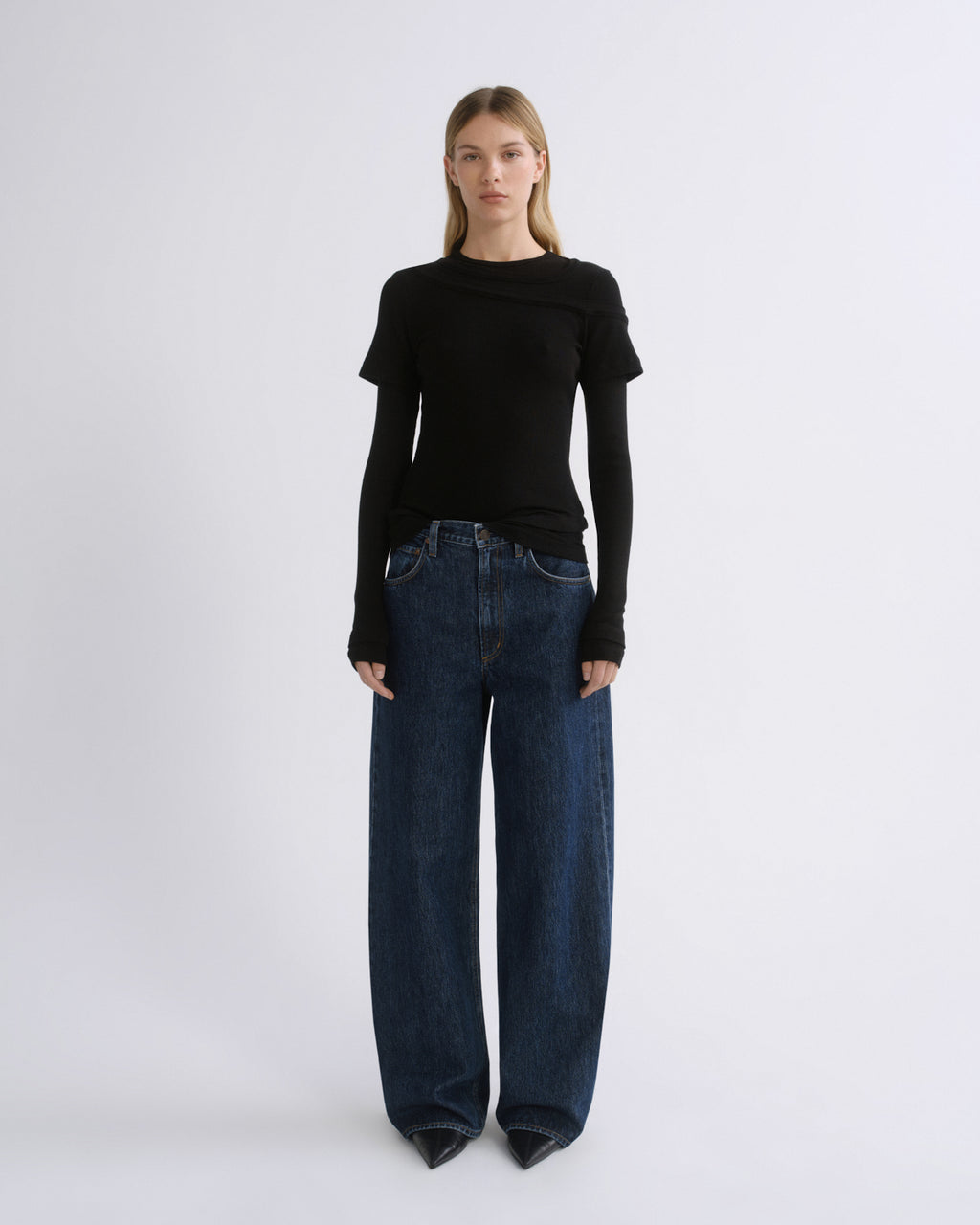 A model stands against a light gray studio background wearing a black long-sleeve top with a draped neckline and dark blue wide-leg jeans.
