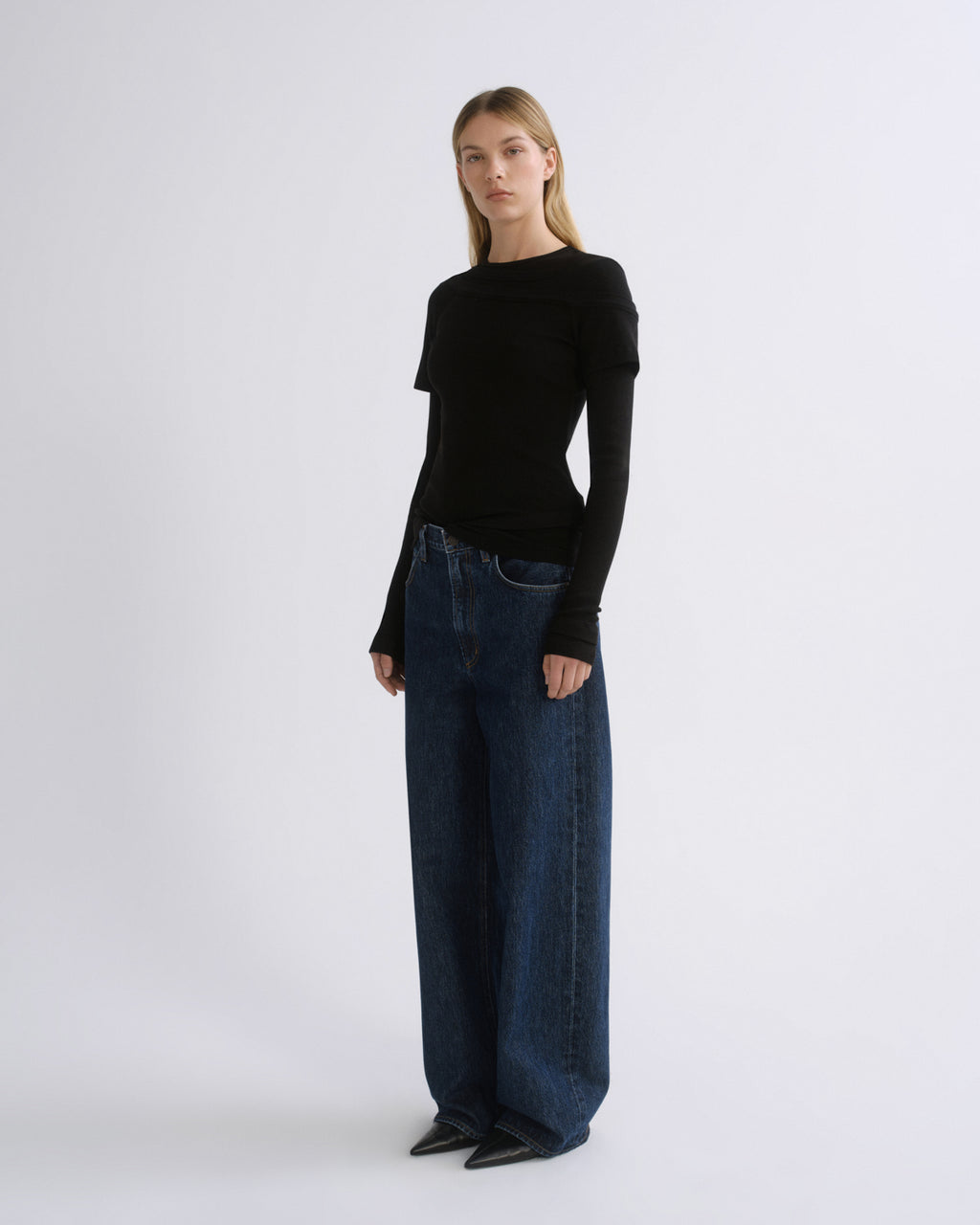 A model stands against a light gray studio background wearing a fitted black long-sleeve top, wide-leg dark blue jeans, and black pointed shoes.