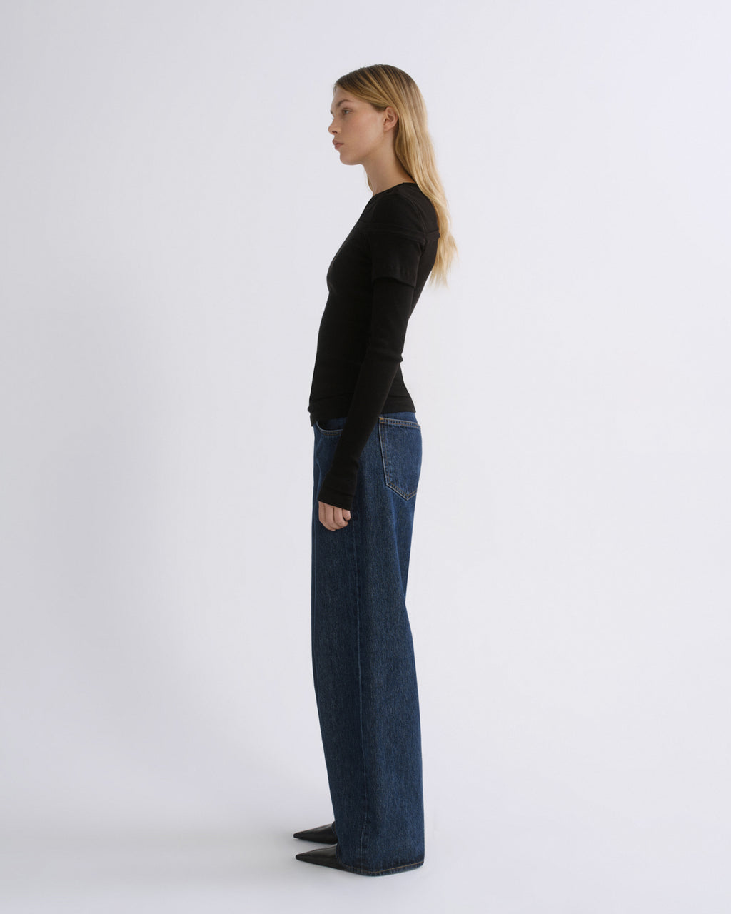 A woman stands in profile wearing a fitted black long-sleeve top and dark blue wide-leg jeans against a plain light gray studio background.