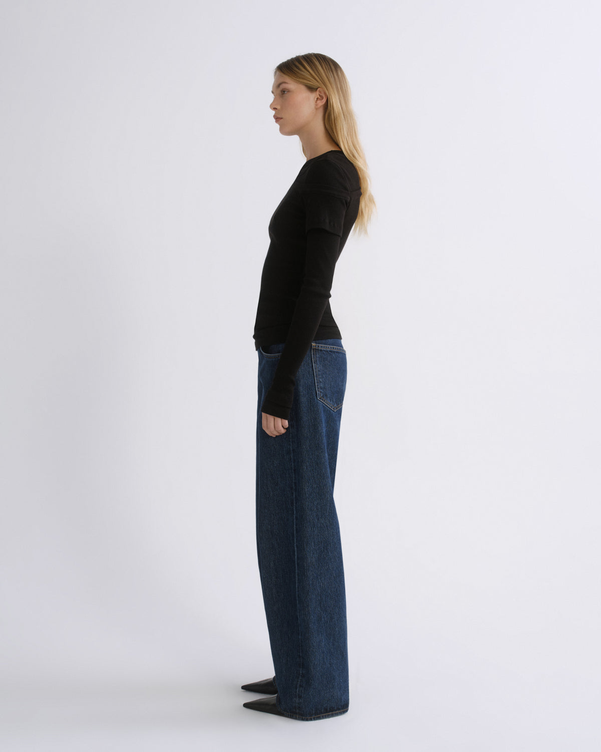A woman stands in profile wearing a fitted black long-sleeve top and dark blue wide-leg jeans against a plain light gray studio background.