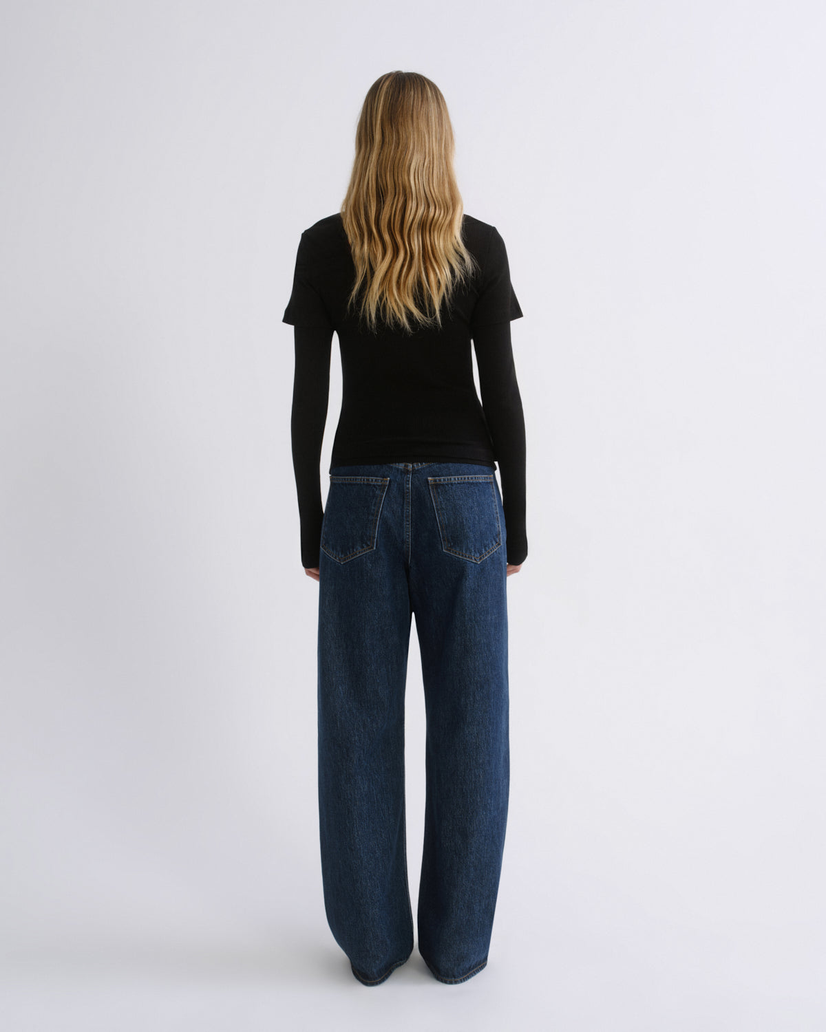 A woman viewed from behind wearing a black long-sleeve top and dark blue wide-leg jeans against a plain light gray studio background.