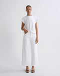 A model stands against a plain light gray studio background wearing a white short-sleeve top tied at the waist, white wide-leg cropped pants, and black flip-flop sandals.