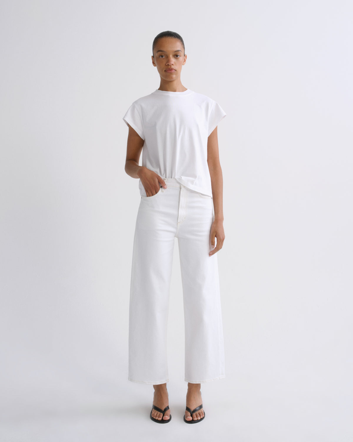 A model stands against a plain light gray studio background wearing a white short-sleeve top tied at the waist, white wide-leg cropped pants, and black flip-flop sandals.