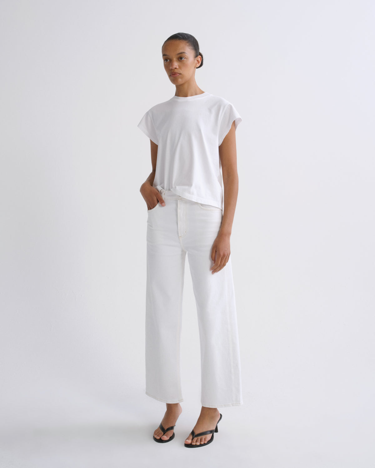 A model stands against a plain light gray studio background wearing a white short-sleeve top and white wide-leg cropped pants, with black flip-flop sandals.