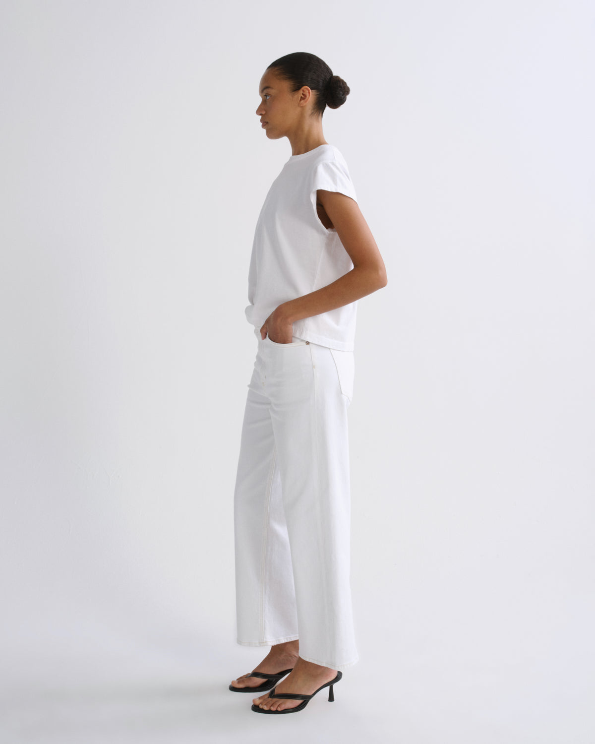 A woman stands in profile wearing a white short-sleeve top and white wide-leg pants with black heeled sandals against a clean white studio background.