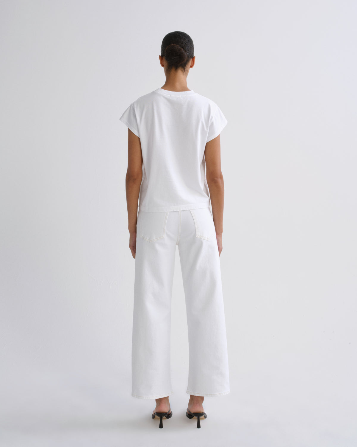A model viewed from the back wearing a white short-sleeve t-shirt and white wide-leg trousers, paired with black high-heeled sandals, against a solid light gray studio background.