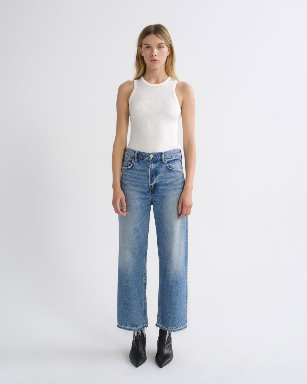 A model stands against a light gray studio background wearing a white sleeveless top, blue wide-leg jeans, and black pointed boots.