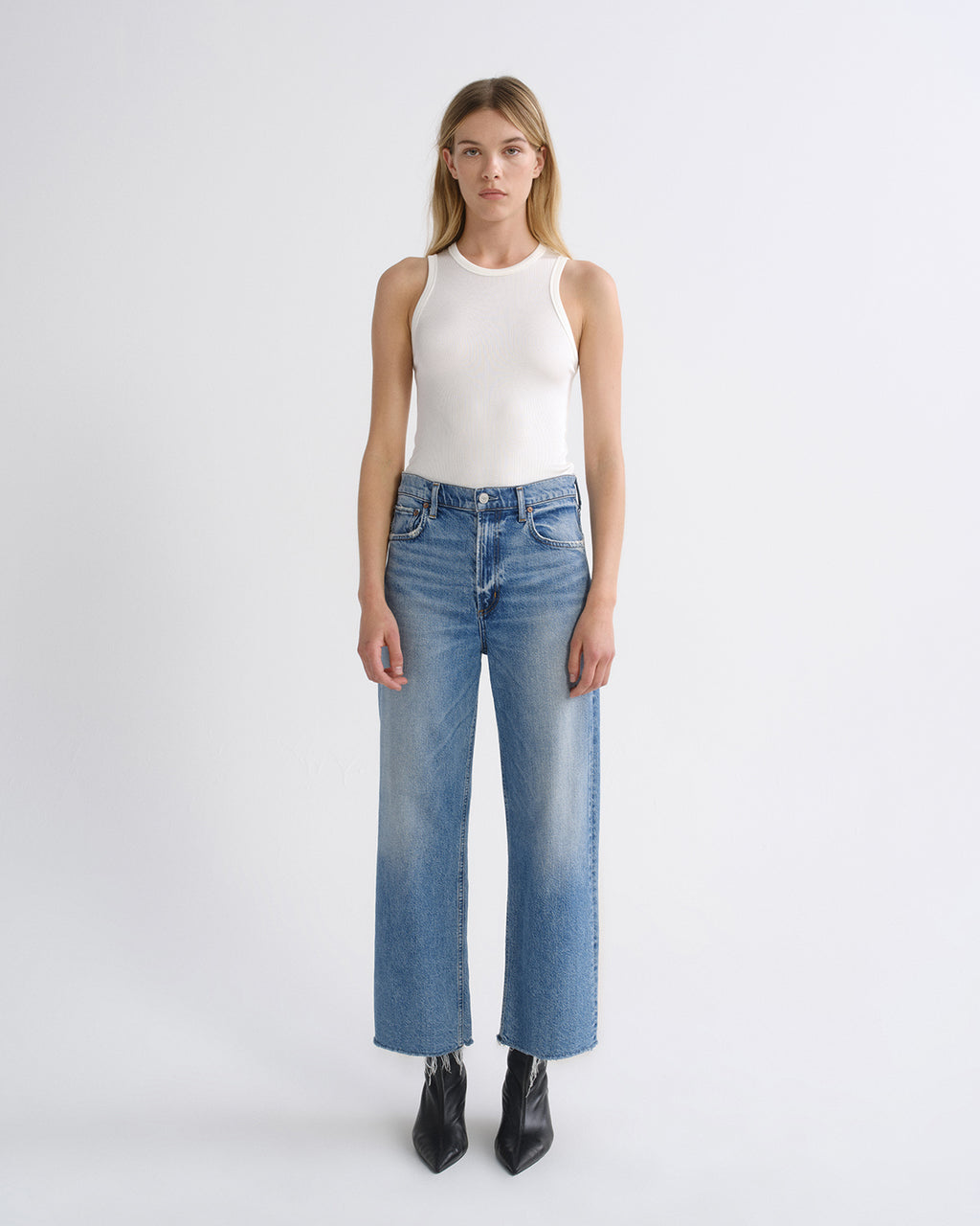 A model stands against a light gray studio background wearing a white sleeveless top, blue wide-leg cropped jeans with a faded wash, and black pointed ankle boots.