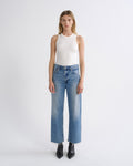 A model stands against a light gray studio background wearing a white sleeveless top, blue wide-leg cropped jeans with a faded wash, and black pointed ankle boots.