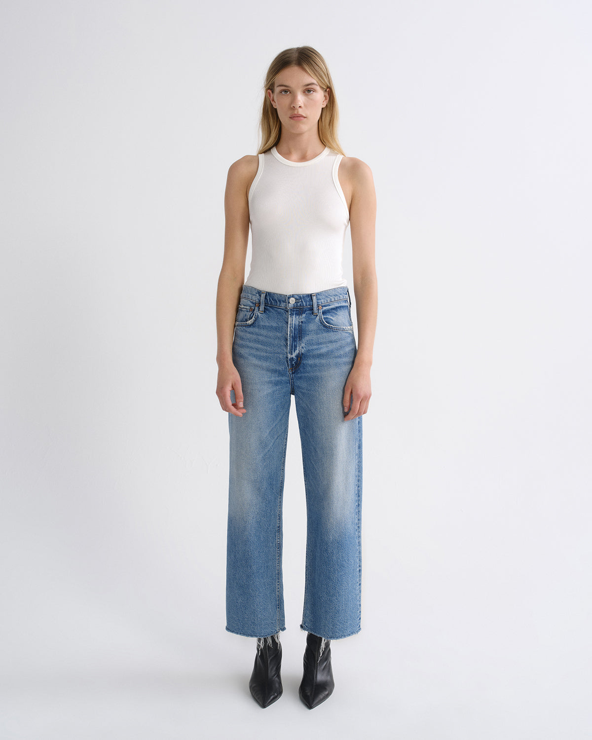 A model stands against a light gray studio background wearing a white sleeveless top, blue wide-leg cropped jeans with a faded wash, and black pointed ankle boots.