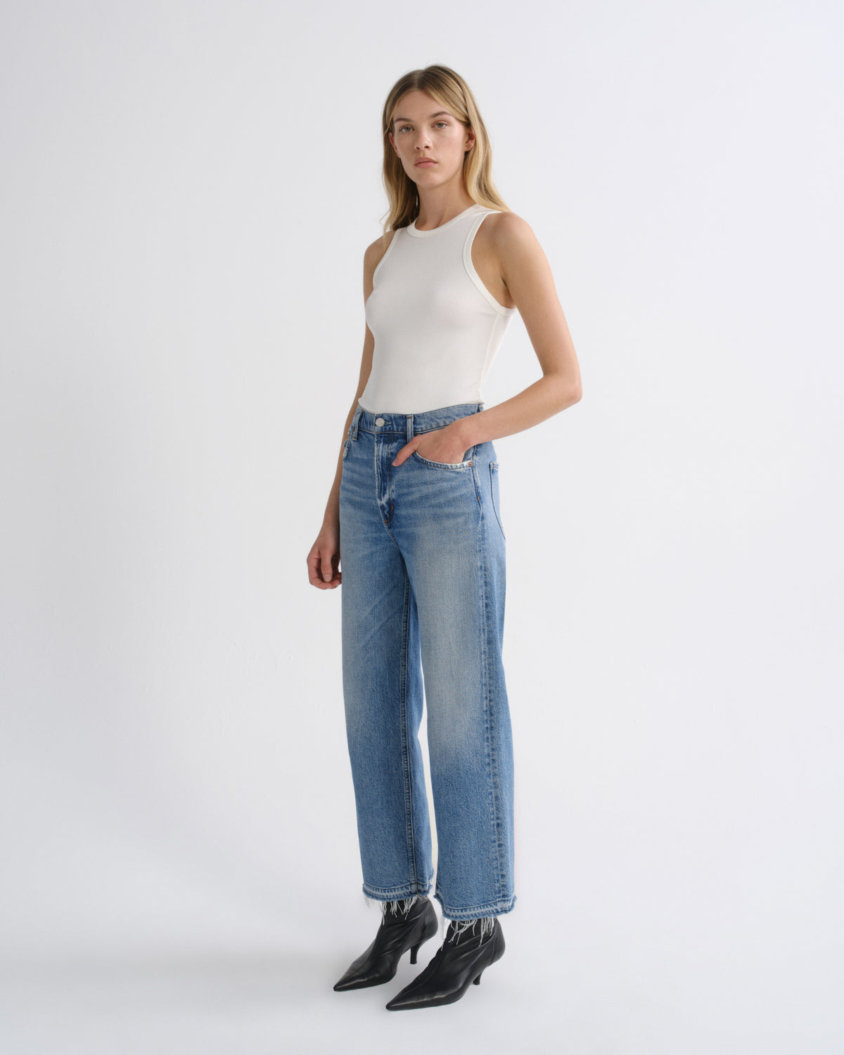 A woman modeling a pair of high-waisted, wide-leg blue jeans with a straight-to-slightly-flared leg, paired with a white sleeveless top and black ankle boots, against a plain light gray studio background.