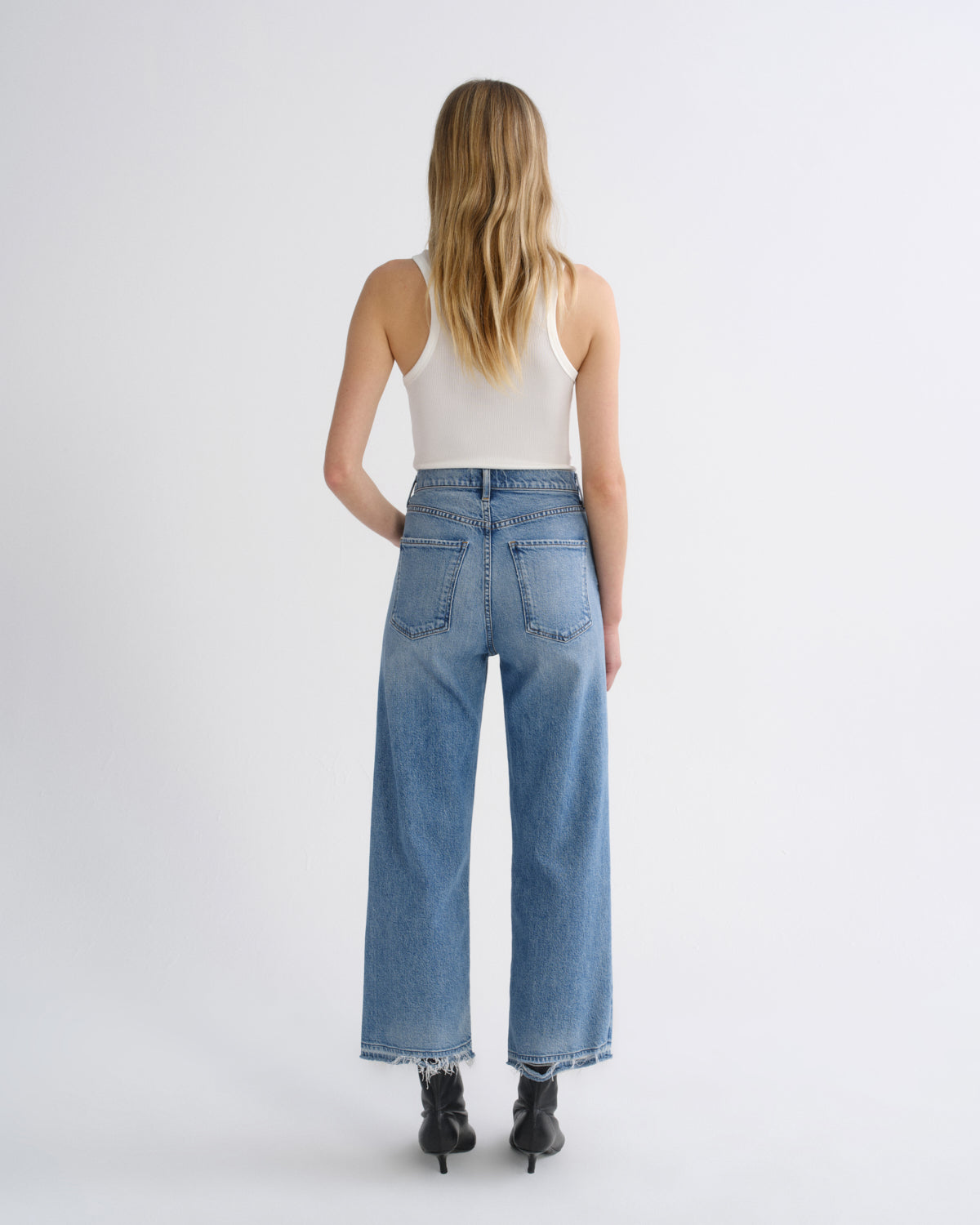 A woman viewed from behind wearing light-wash wide-leg blue jeans, a white ribbed tank top, and black ankle boots against a plain light gray studio background.