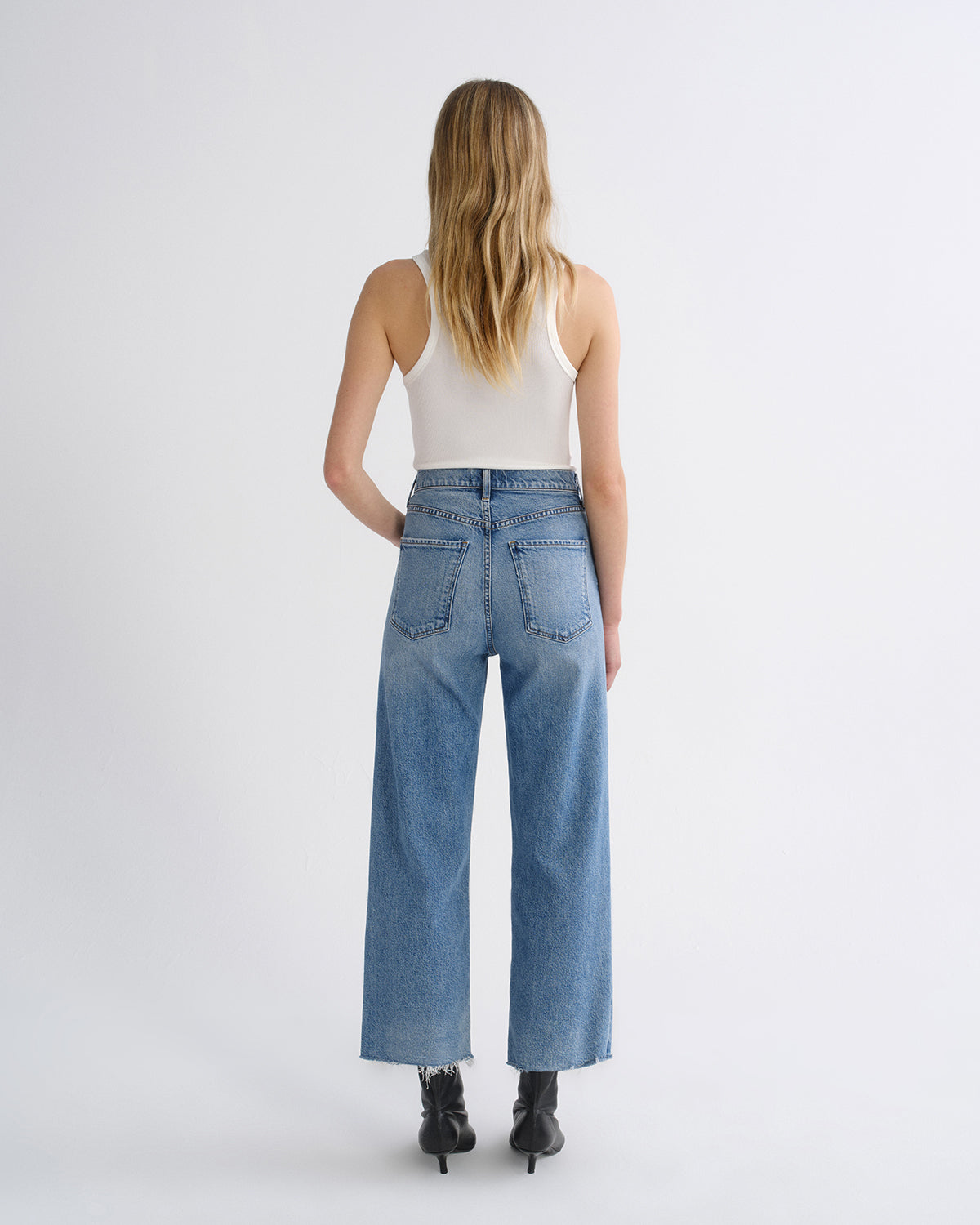 A woman viewed from behind wearing light-wash wide-leg blue jeans, a white ribbed tank top, and black ankle boots against a plain light gray studio background.