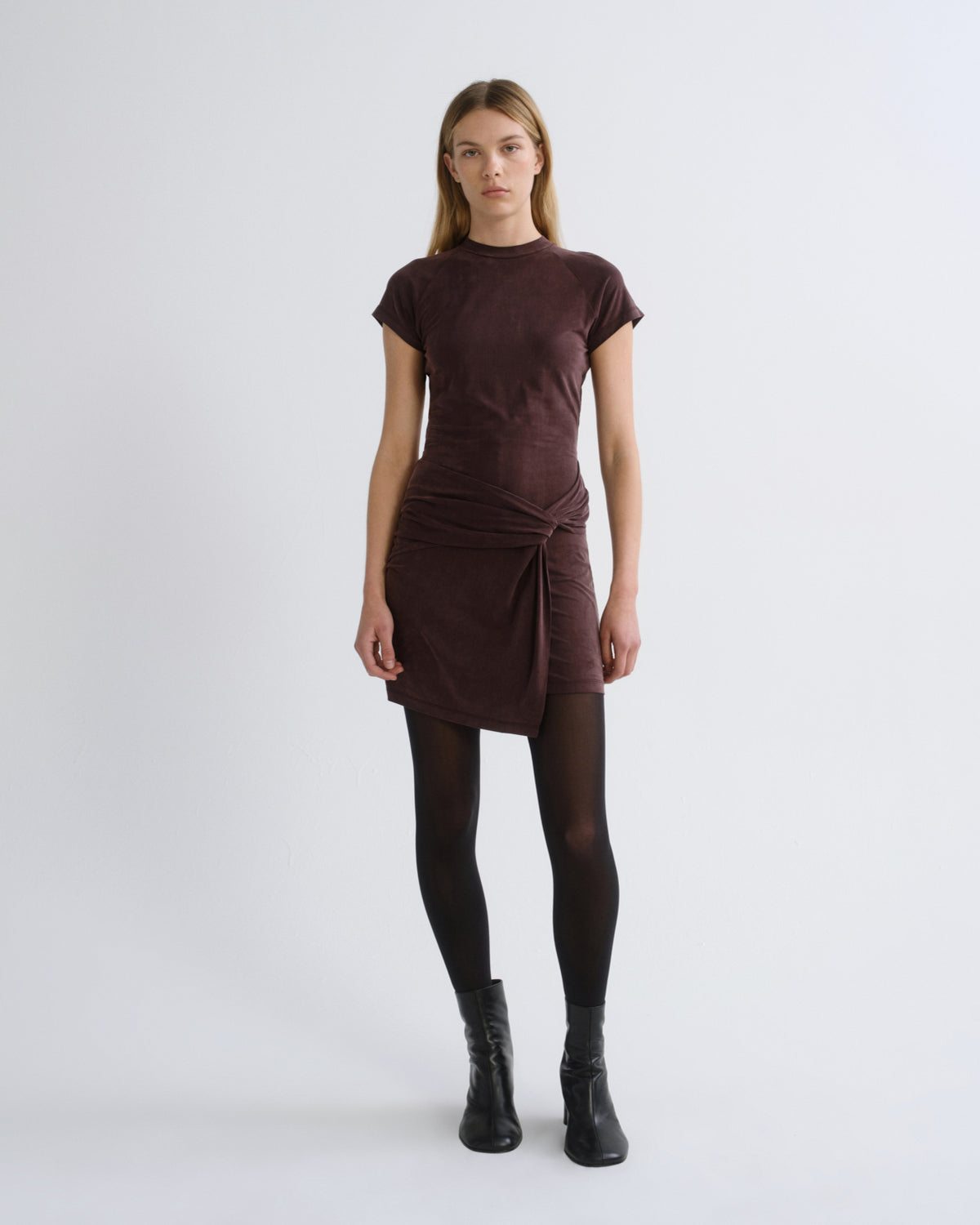 Woman wearing a dark brown short sleeve mini dress with black tights and black boots on a grey background.