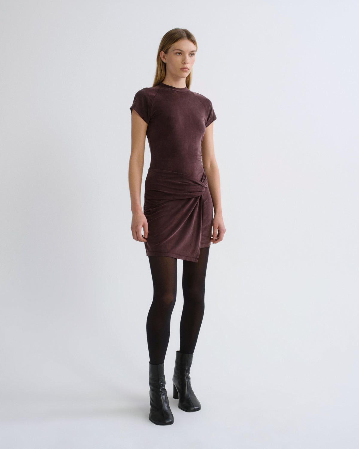 Woman wearing a dark brown short sleeve mini dress with black tights and black boots on a grey background.