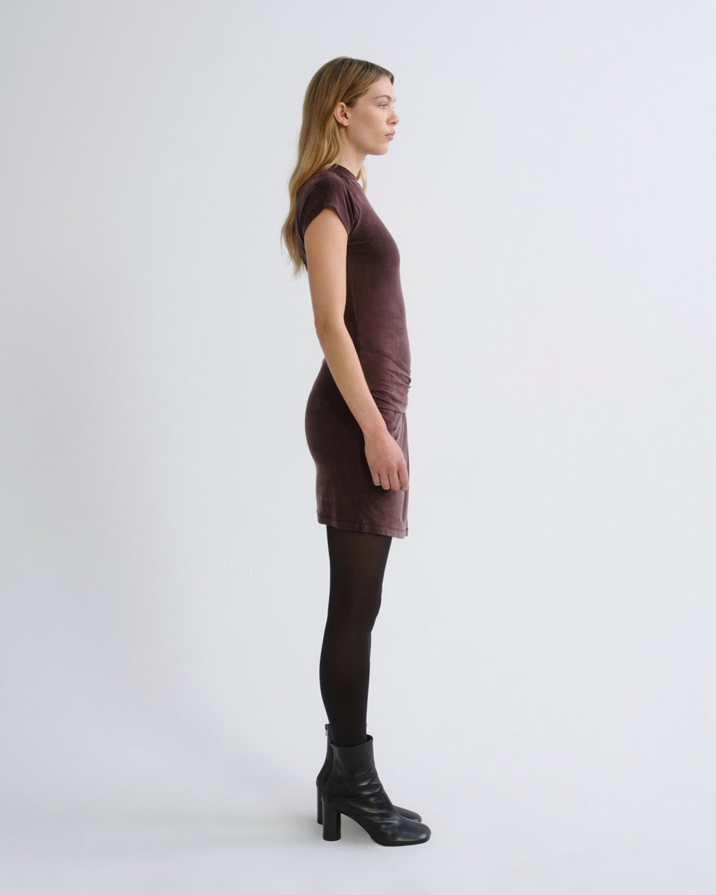 Woman wearing a dark brown short sleeve mini dress with black tights and black boots viewed from the side on a grey background.
