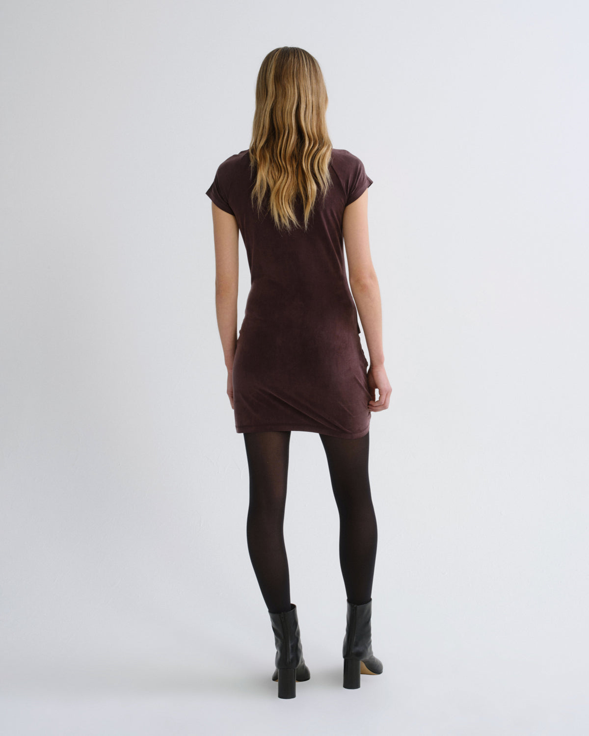 A woman viewed from behind wearing a short-sleeve dark plum velvet mini dress, black tights, and black ankle boots against a plain light gray studio background.