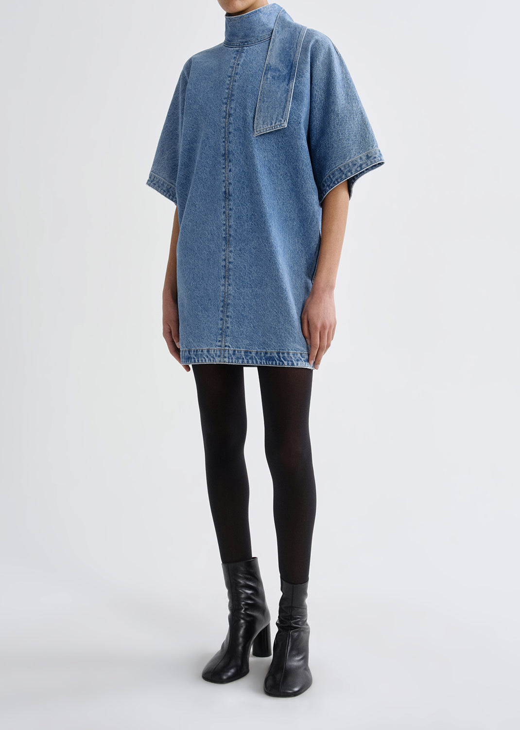 Person wearing a blue denim dress over black leggings on a grey background