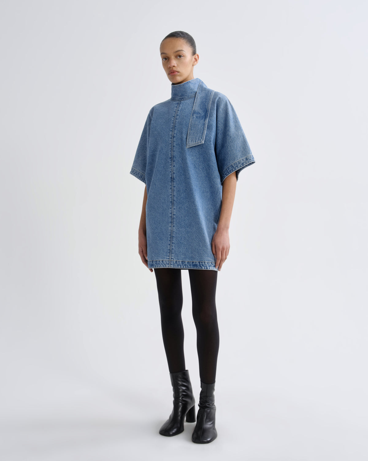A model stands against a light gray studio background wearing a light blue denim tunic dress with a high mock neck, short wide sleeves, and a front patch pocket. The dress has raw, unfinished hems and is styled with black tights and black leather ankle boots.