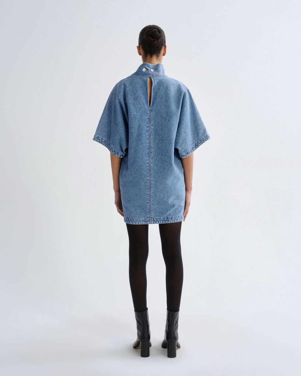 A model viewed from the back wearing a light blue denim tunic top with short wide sleeves, a high collar, and a central back slit, styled with black tights and black boots against a light gray studio background.
