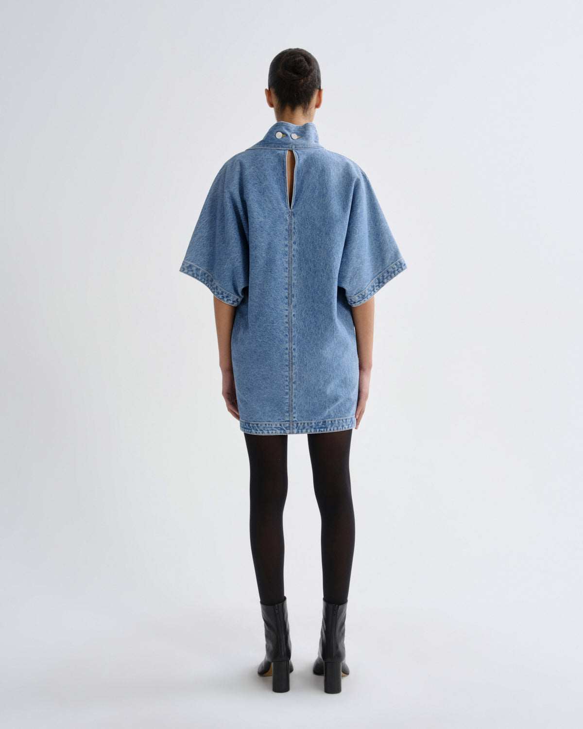 A model viewed from the back wearing a light blue denim tunic top with short wide sleeves, a high collar, and a central back slit, styled with black tights and black boots against a light gray studio background.
