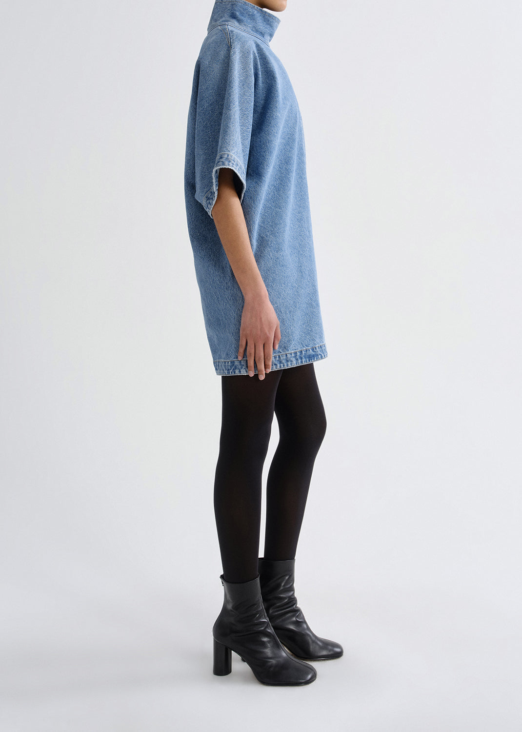Person wearing a blue denim dress with black tights and boots on a grey background