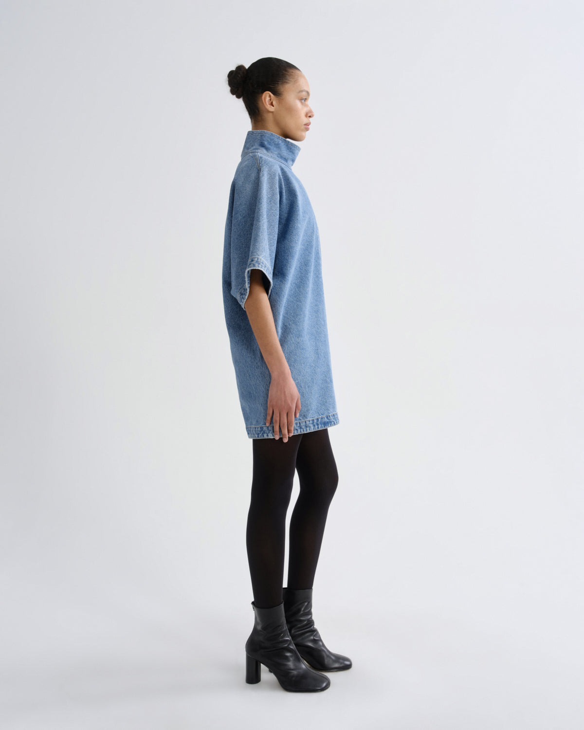 A model in profile wearing a light blue denim tunic dress with short wide sleeves, black tights, and black leather ankle boots, standing against a plain light gray studio background.
