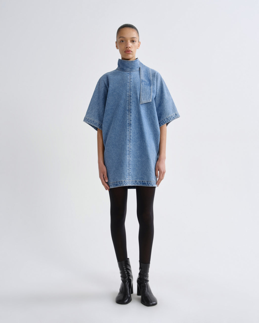 A model stands against a light gray studio background wearing a light blue denim short-sleeve shirt dress with a high mock neck, black tights, and black ankle boots.