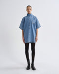 A model stands against a light gray studio background wearing a light blue denim short-sleeve shirt dress with a high mock neck, black tights, and black ankle boots.
