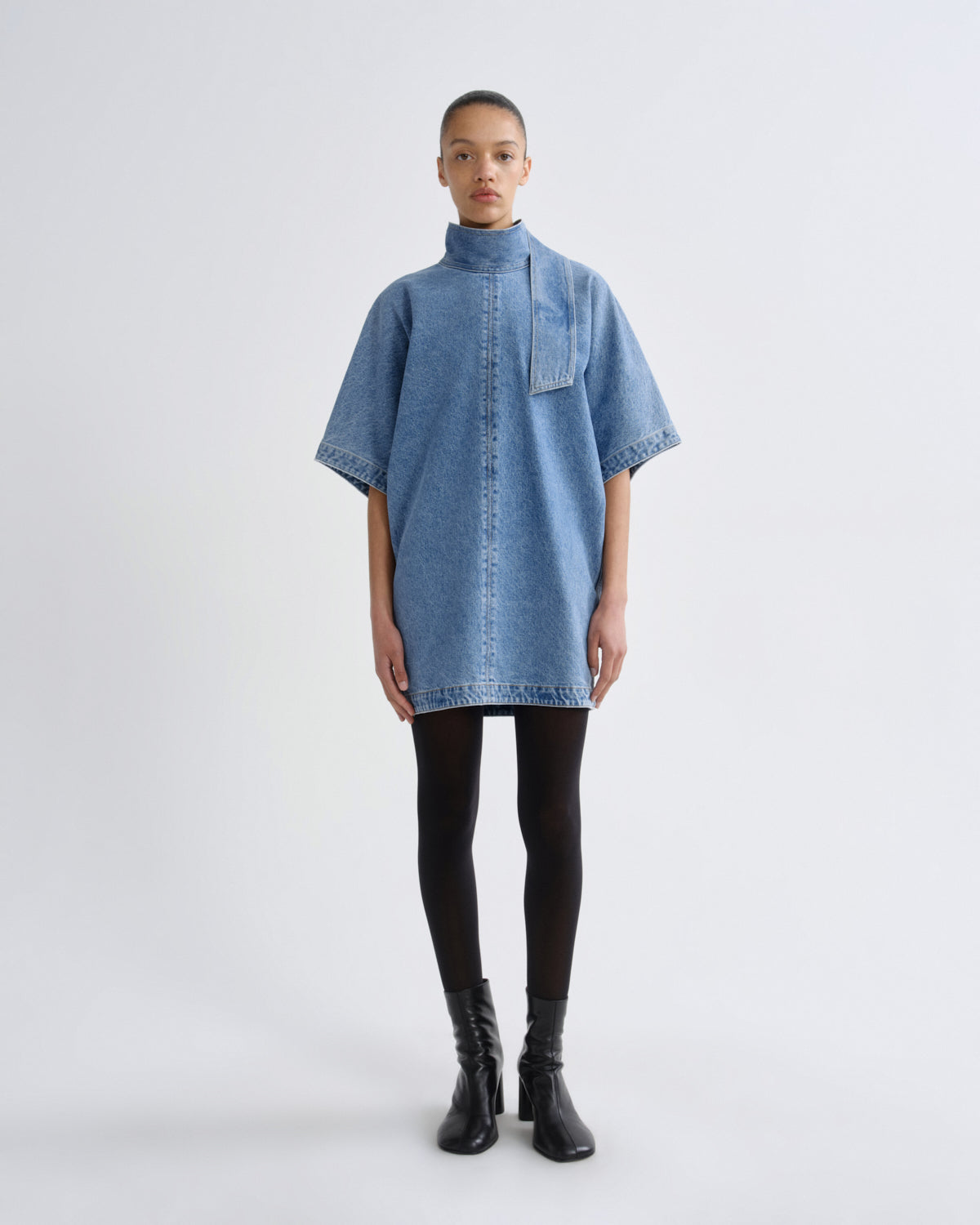 A model stands against a light gray studio background wearing a light blue denim short-sleeve shirt dress with a high mock neck, black tights, and black ankle boots.